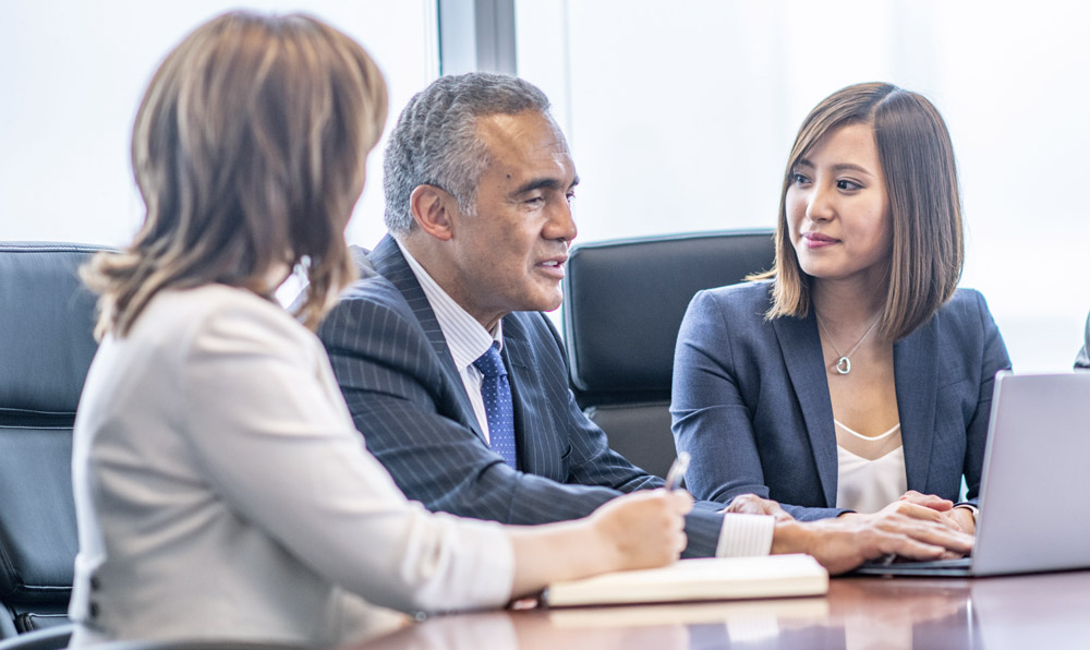 A group of young businesspeople are indoors in a board meeting room. They are sitting and having a discussion about Opus 2
